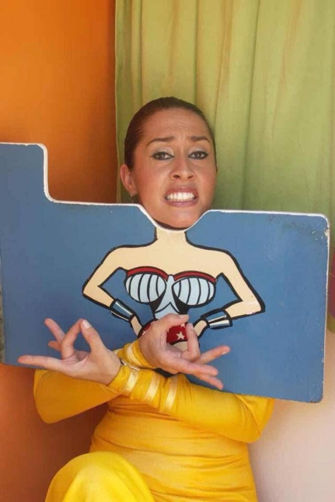 CarOlé Flamenco in a yellow outfit holding a painted strongwoman board against an orange wall